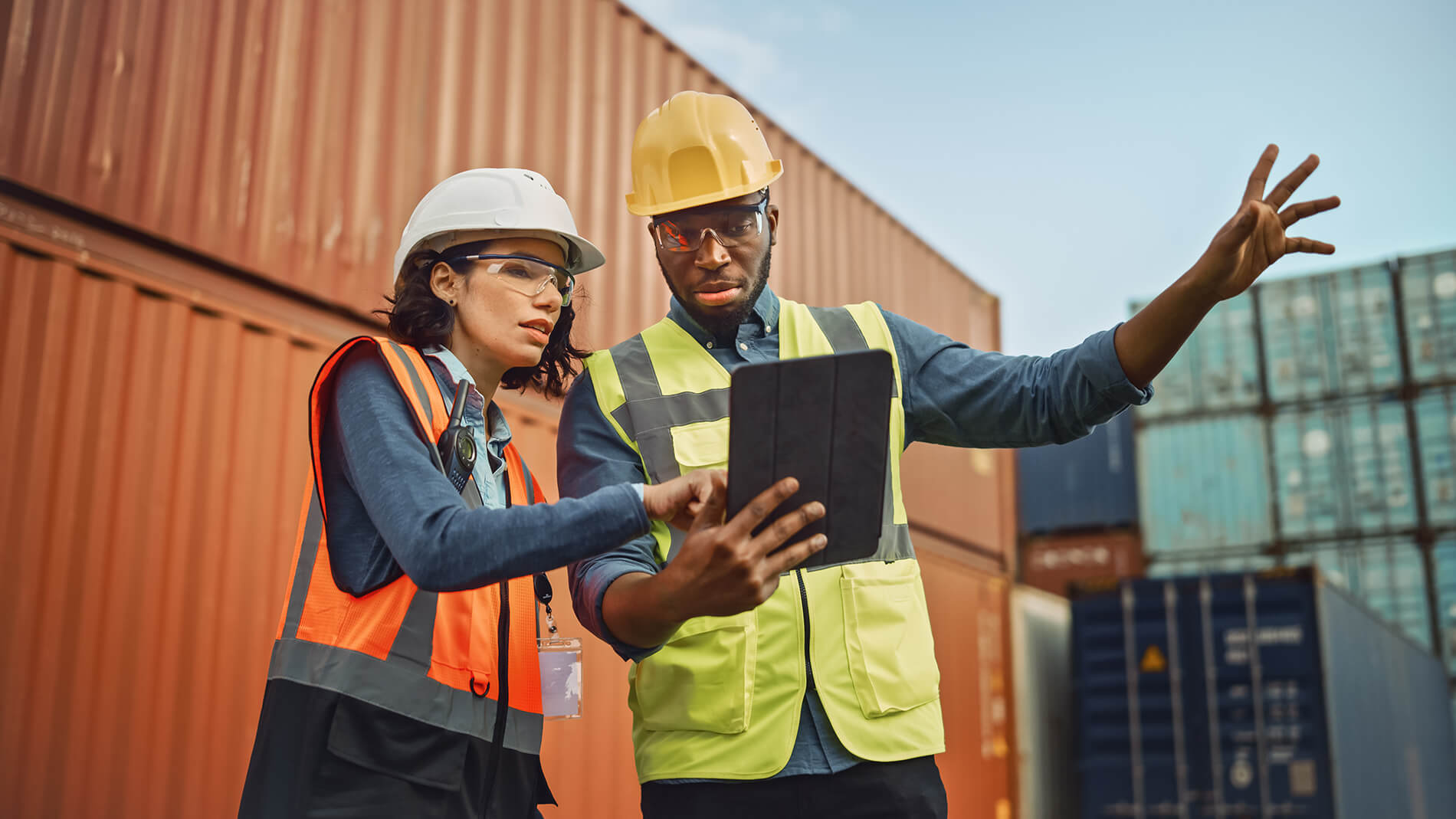 facilities-staff-recruitment A woman and a man looking at a tablet with an industrial setting of shipping containers behind them